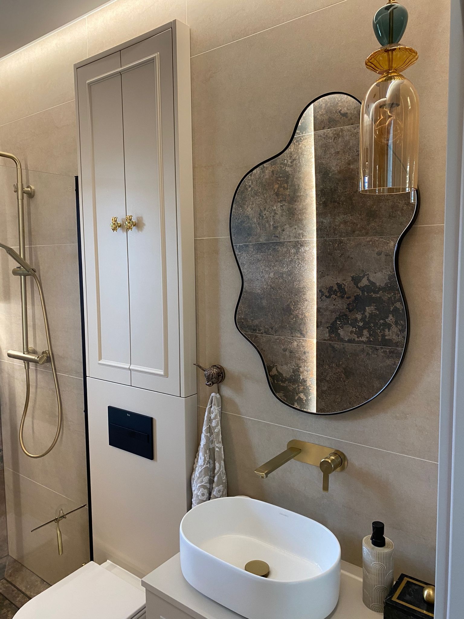 Elegancka łazienka w beżach: asymetryczne lustro, złota armatura, kabina prysznicowa i szafka z dekoracyjnymi uchwytami. Subtelne oświetlenie podkreśla detale.