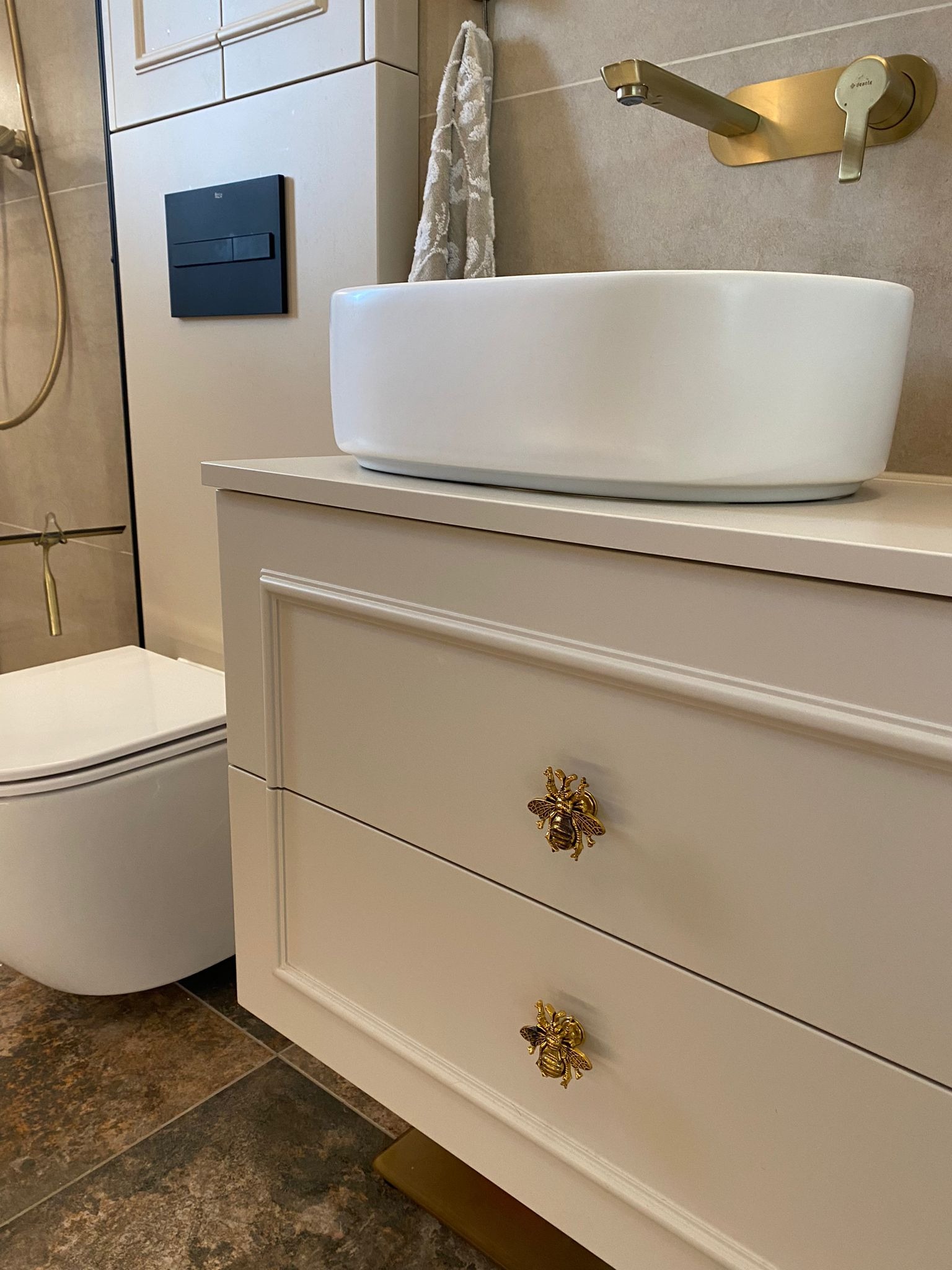 Beżowa szafka łazienkowa z umywalką nablatową i złotą armaturą. Uwagę przykuwają ozdobne uchwyty w kształcie pszczół. Widoczny fragment toalety i brązowa podłoga.