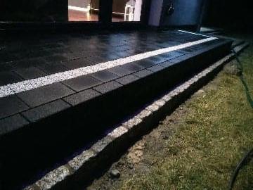 Ciemna kostka brukowa ułożona na schodach zewnętrznych budynku, oddzielona białym pasem z drobnego kamienia, widoczne oświetlenie budynku w tle.