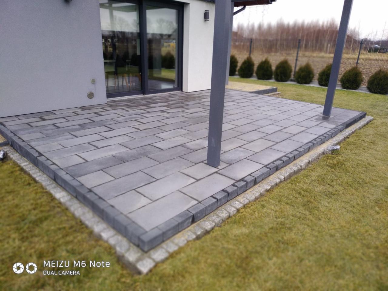 Nowoczesny taras z szarej kostki brukowej w kształcie prostokątów, obrzeża z kostki granitowej, widok na przesuwne drzwi tarasowe i metalowe słupy zadaszenia, trawnik wokół.