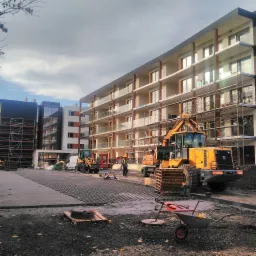 Budowa nowego budynku wielorodzinnego w Opolu, z widocznymi rusztowaniami, ciężkim sprzętem budowlanym i pracownikami w żółtych kaskach.