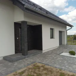 Nowoczesny dom jednorodzinny z elewacją w kolorze złamanej bieli, z fragmentami wykończonymi czarnym kamieniem dekoracyjnym przy wejściu i garażu, oraz szarą kostką brukową na podjeździe i ścieżkach.