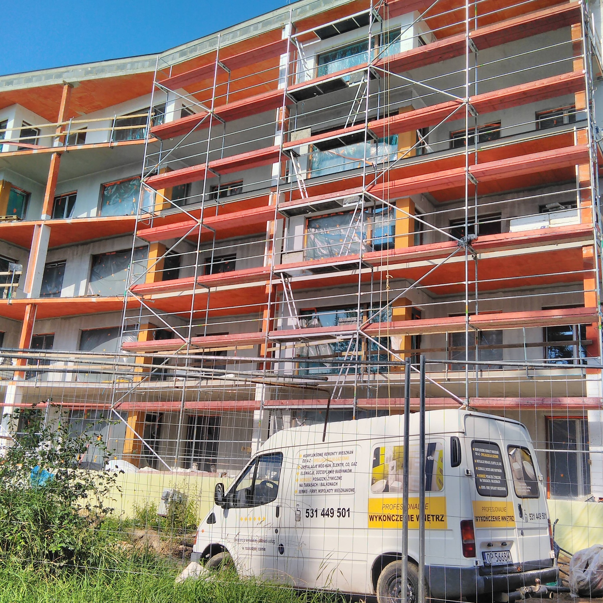 Budynek wielorodzinny w trakcie prac elewacyjnych, widoczne rusztowania, zabezpieczone okna folią, na pierwszym planie samochód dostawczy z reklamą usług remontowych i wykończeniowych.