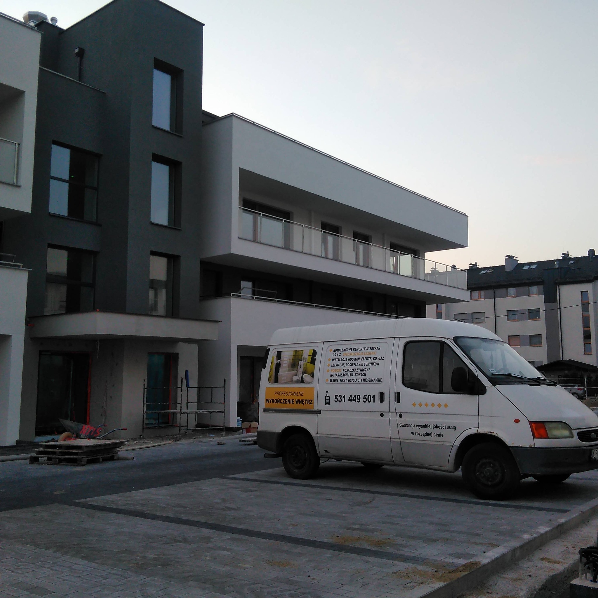 Nowoczesny budynek mieszkalny z szarą i białą elewacją, częściowo w trakcie wykończenia, z zaparkowanym białym vanem z reklamą firmy remontowej z hasłem 'Profesjonalne wykończenie wnętrz' i numerem...