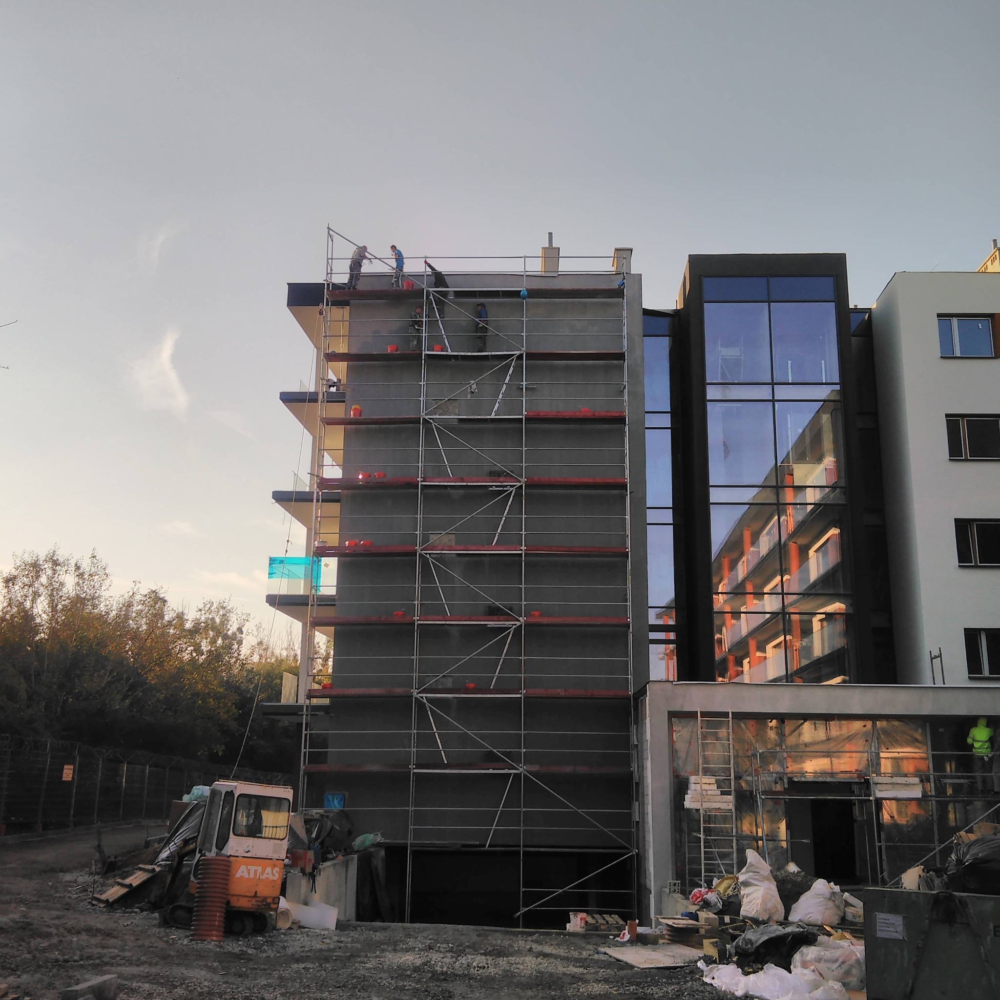 Wielopiętrowy budynek w trakcie prac elewacyjnych; widoczne rusztowania, pracownicy oraz sprzęt budowlany, w tle drzewa i zachodzące słońce.