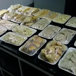 Różnorodne dania, w tym pierogi, kluski i ciasta, zapakowane w plastikowe tacki i folie, ułożone rzędami na czarnym blacie, gotowe do transportu.