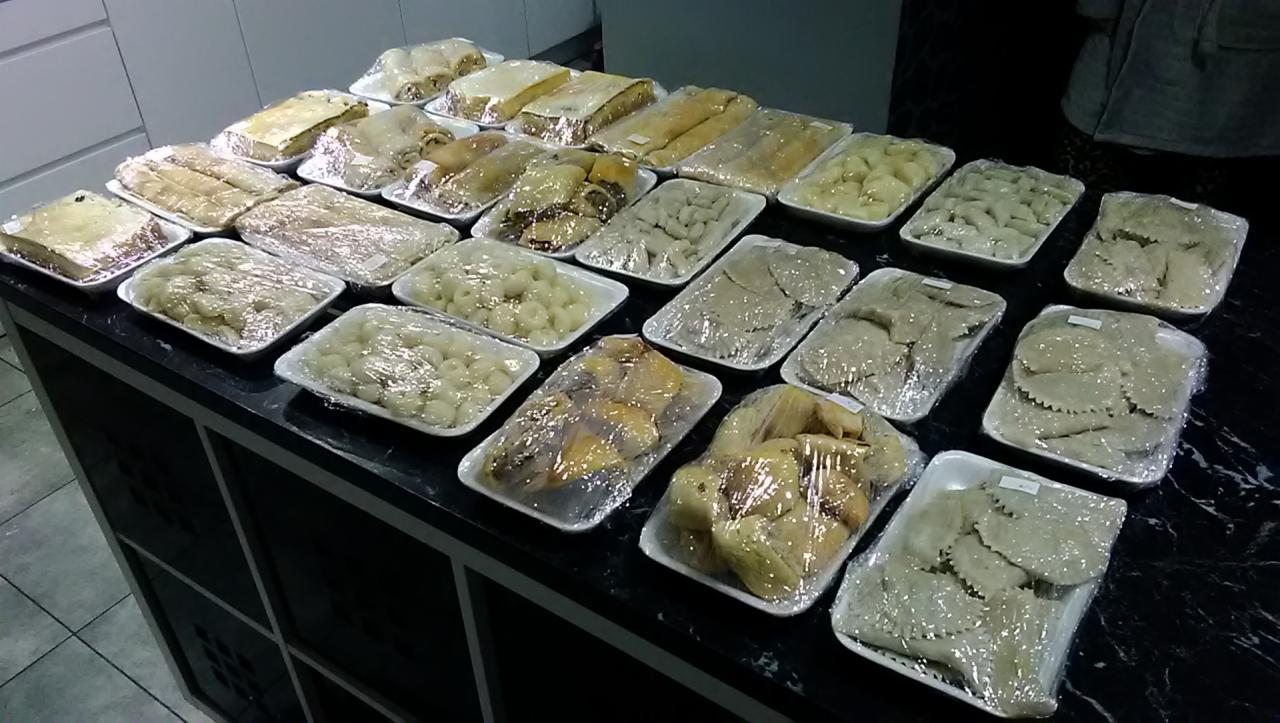 Różnorodne dania, w tym pierogi, kluski i ciasta, zapakowane w plastikowe tacki i folie, ułożone rzędami na czarnym blacie, gotowe do transportu.