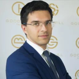 Portret mężczyzny w okularach i granatowym garniturze na tle z powtarzającym się złotym logo firmy.