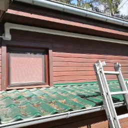 Rynna z tworzywa sztucznego zamontowana na drewnianym domu, drabina oparta o dach z zielonej blachodachówki, widoczne igliwie na dachu.