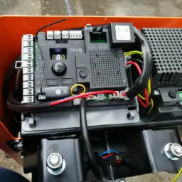 Otwarta obudowa sterownika Nice z widocznymi elementami elektroniki, w tym wyświetlaczem, złączami i przewodami, zainstalowanego wewnątrz pomarańczowej obudowy.