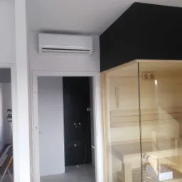 Biały klimatyzator ścienny zamontowany w nowoczesnym wnętrzu z sauną, widoczne niedokończone prace wykończeniowe, w tle czarna ściana.