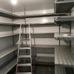 Wnętrze chłodni z metalowymi, perforowanymi półkami i rozstawioną drabiną aluminiową z czerwonymi akcentami na stopniach.