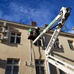 Pracownik w zielonej kurtce na podnośniku koszowym, serwisujący klimatyzatory na ścianie budynku w Kolsku. Widoczny numer telefonu na ramieniu podnośnika.