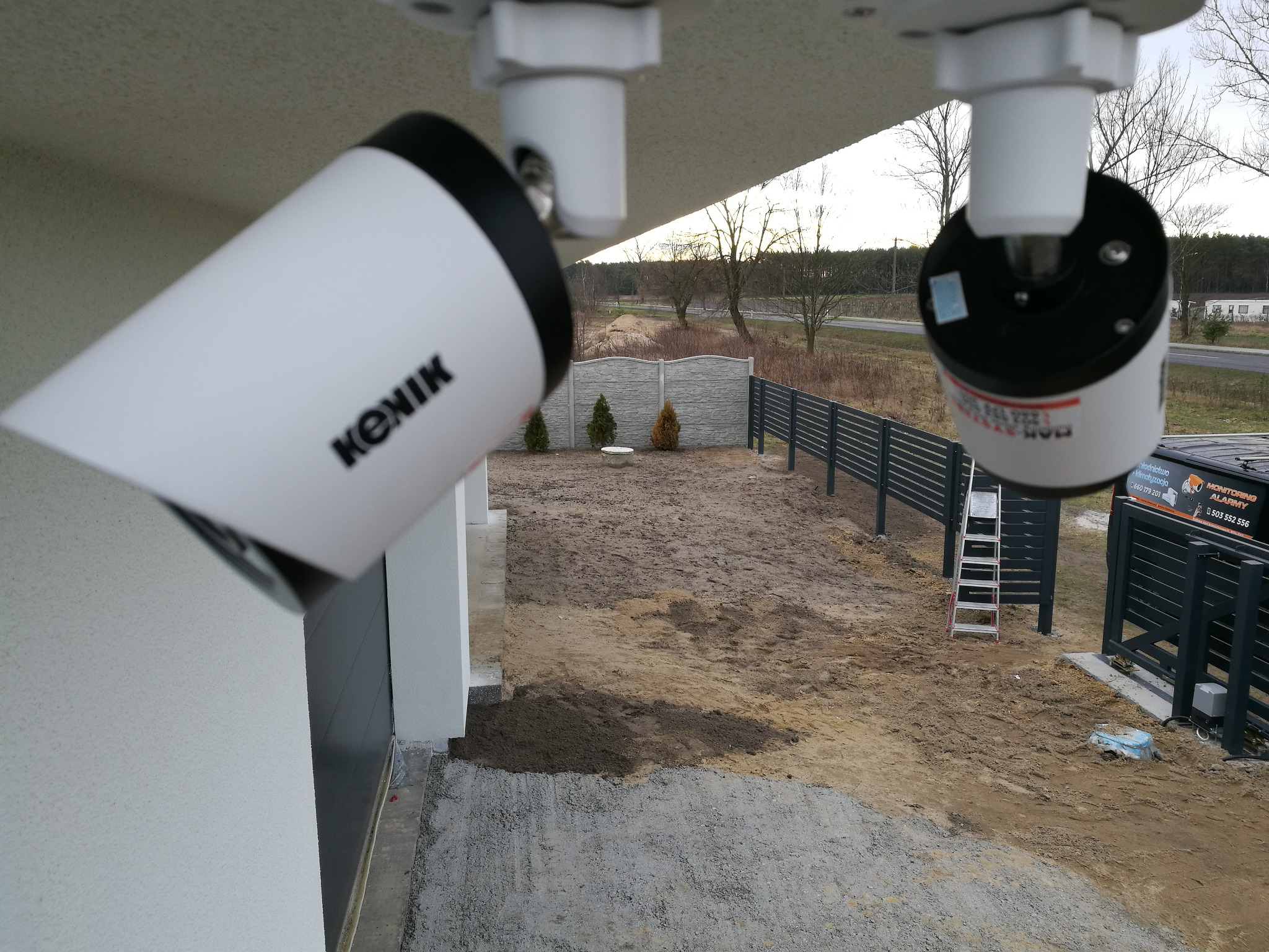 Dwie kamery monitoringu marki KONIK zamontowane pod okapem budynku, widok na posesję z szarą, betonową podmurówką i metalowym ogrodzeniem, w tle drzewa.
