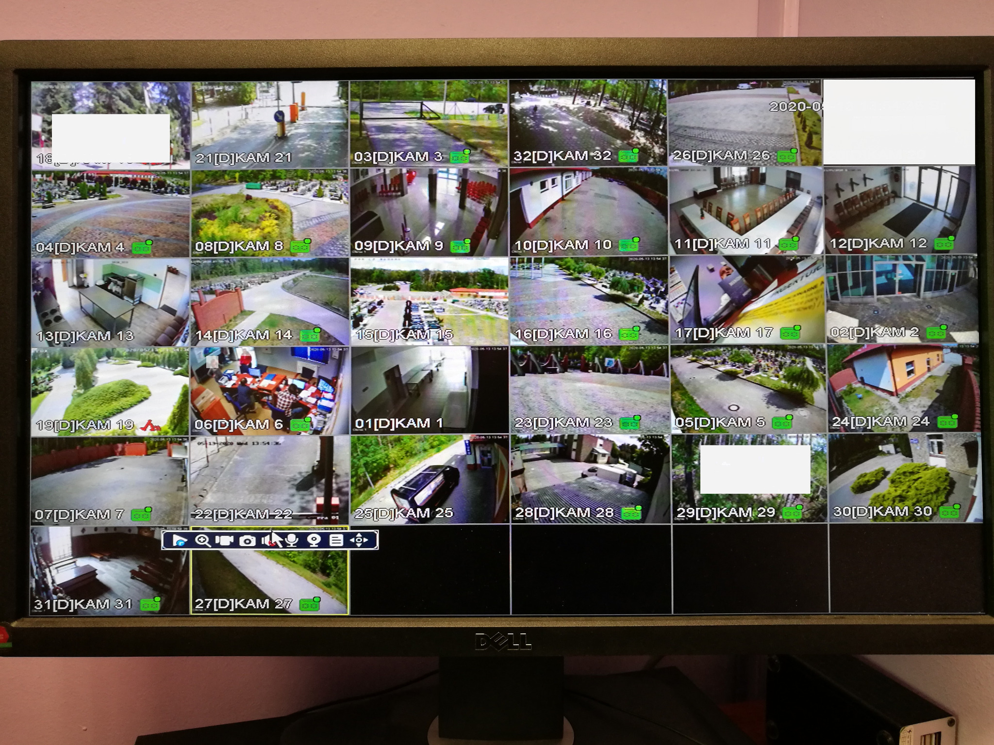 Ekran monitora z podglądem z wielu kamer monitoringu, widoczne różne ujęcia z zewnątrz i wewnątrz budynków, z datami i oznaczeniami kamer.