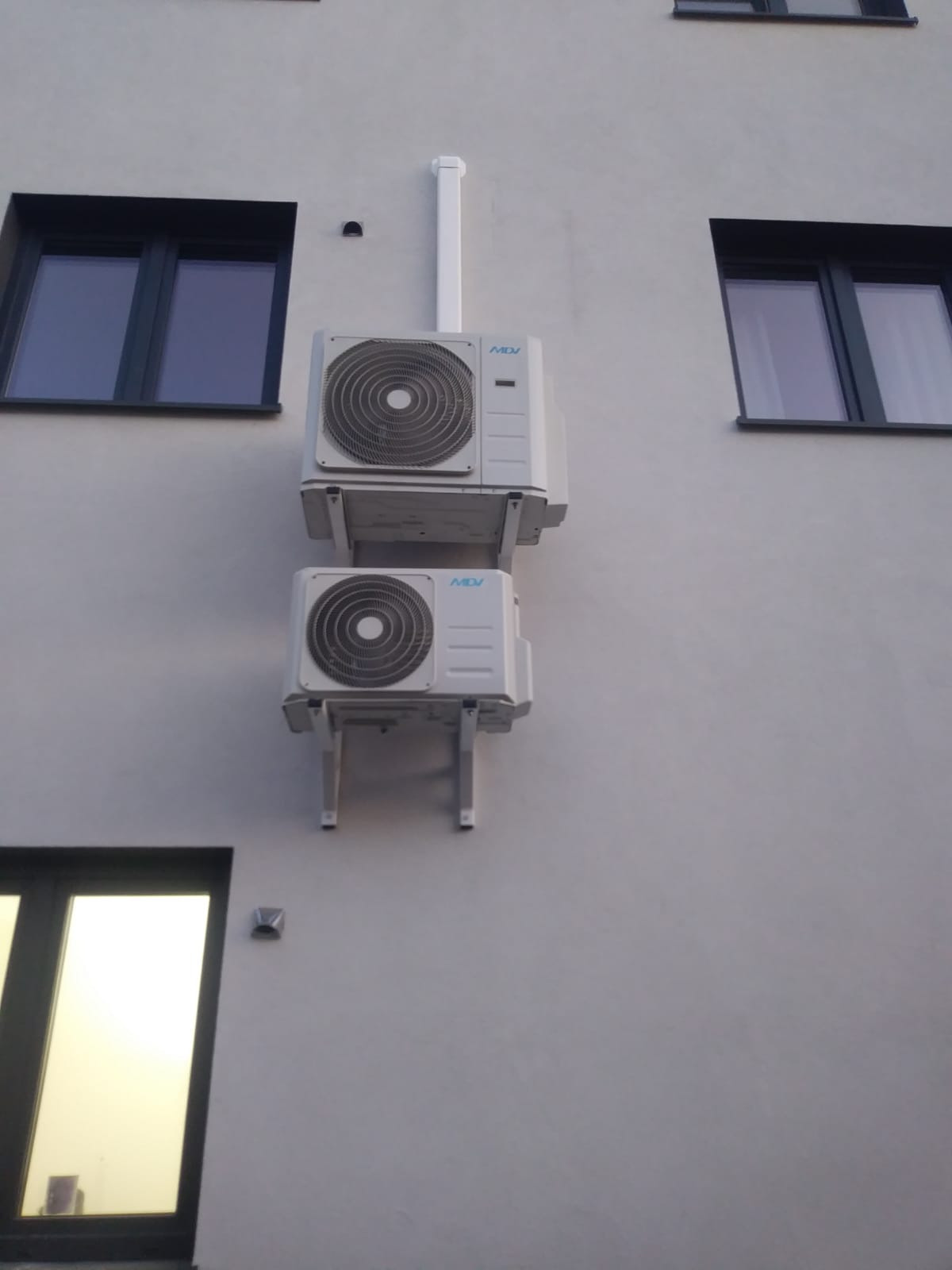Dwa klimatyzatory MDI zamontowane jeden nad drugim na szarej elewacji budynku, widoczne okna z ciemnymi ramami i biała rura odprowadzająca skropliny.