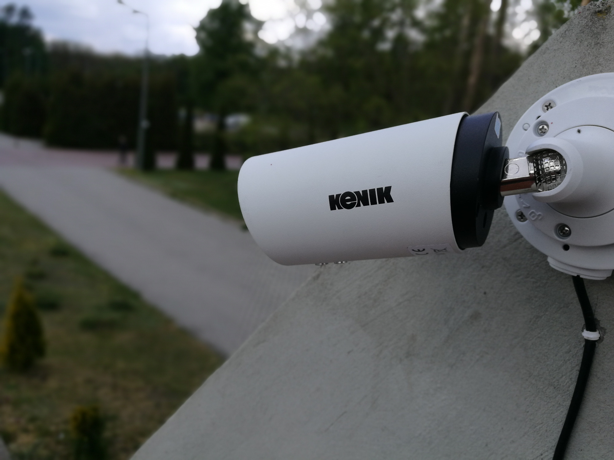 Biała kamera monitoringu marki KENIK zamontowana na szarej ścianie zewnętrznej budynku, widok z boku, z kablem zasilającym i tłem z zielenią parkową.