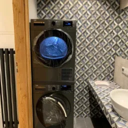 Zestaw pralko-suszarki Beko w nowoczesnej łazience z geometryczną mozaiką na ścianie, jeszcze w folii ochronnej po instalacji.