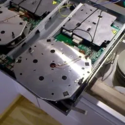 Wnętrze rozmontowanej płyty indukcyjnej z widocznymi elementami elektronicznymi i metalowymi osłonami na blacie kuchennym z jasnego drewna.