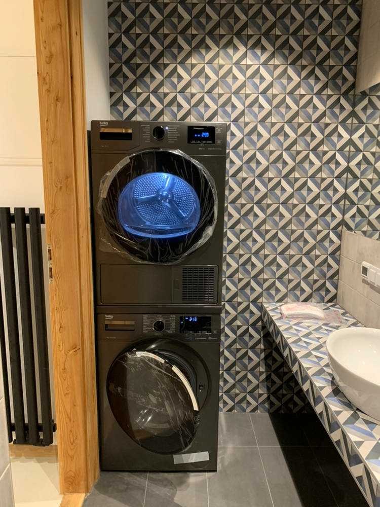 Zestaw pralko-suszarki Beko w nowoczesnej łazience z geometryczną mozaiką na ścianie, jeszcze w folii ochronnej po instalacji.