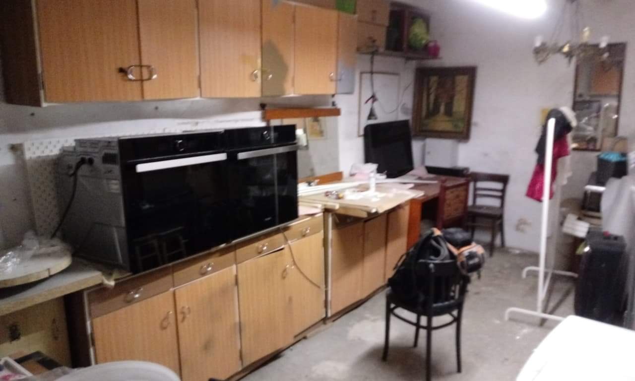 Czarny piekarnik elektryczny w zabudowie kuchennej z drewnianymi szafkami, widoczne podłączenie elektryczne, tło zagracone.