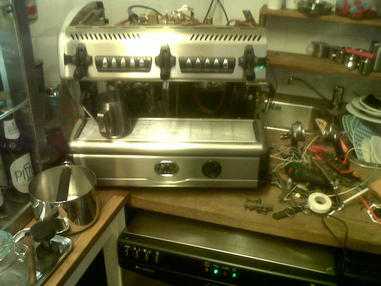 Otwarta, srebrna, profesjonalna maszyna do kawy w trakcie naprawy, z widocznymi przewodami i narzędziami na blacie, w tle butelki i naczynia.