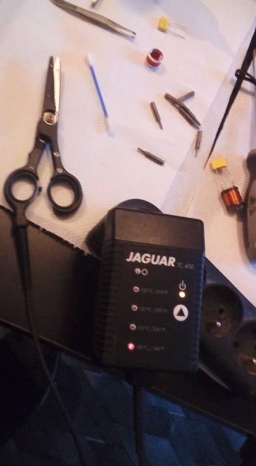 Stół roboczy z rozłożonymi narzędziami i elementami elektronicznymi, w tym czarne nożyczki, pęseta, wacik, kondensatory, wiertła i urządzenie Jaguar TC-400 z diodami LED wskazującymi temperaturę.