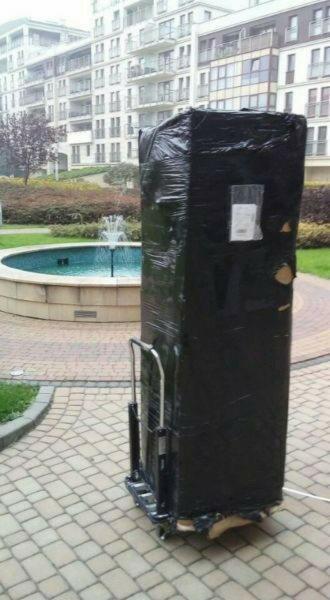Sprzęt AGD (lodówka lub pralka) owinięty folią transportową, umieszczony na wózku transportowym, na tle fontanny i budynku mieszkalnego.