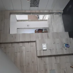 Wnętrze łazienki w trakcie prac wykończeniowych: ułożone płytki imitujące drewno na ścianie i czarne na podłodze, widoczne elementy instalacji wodnej i akcesoria glazurnicze.