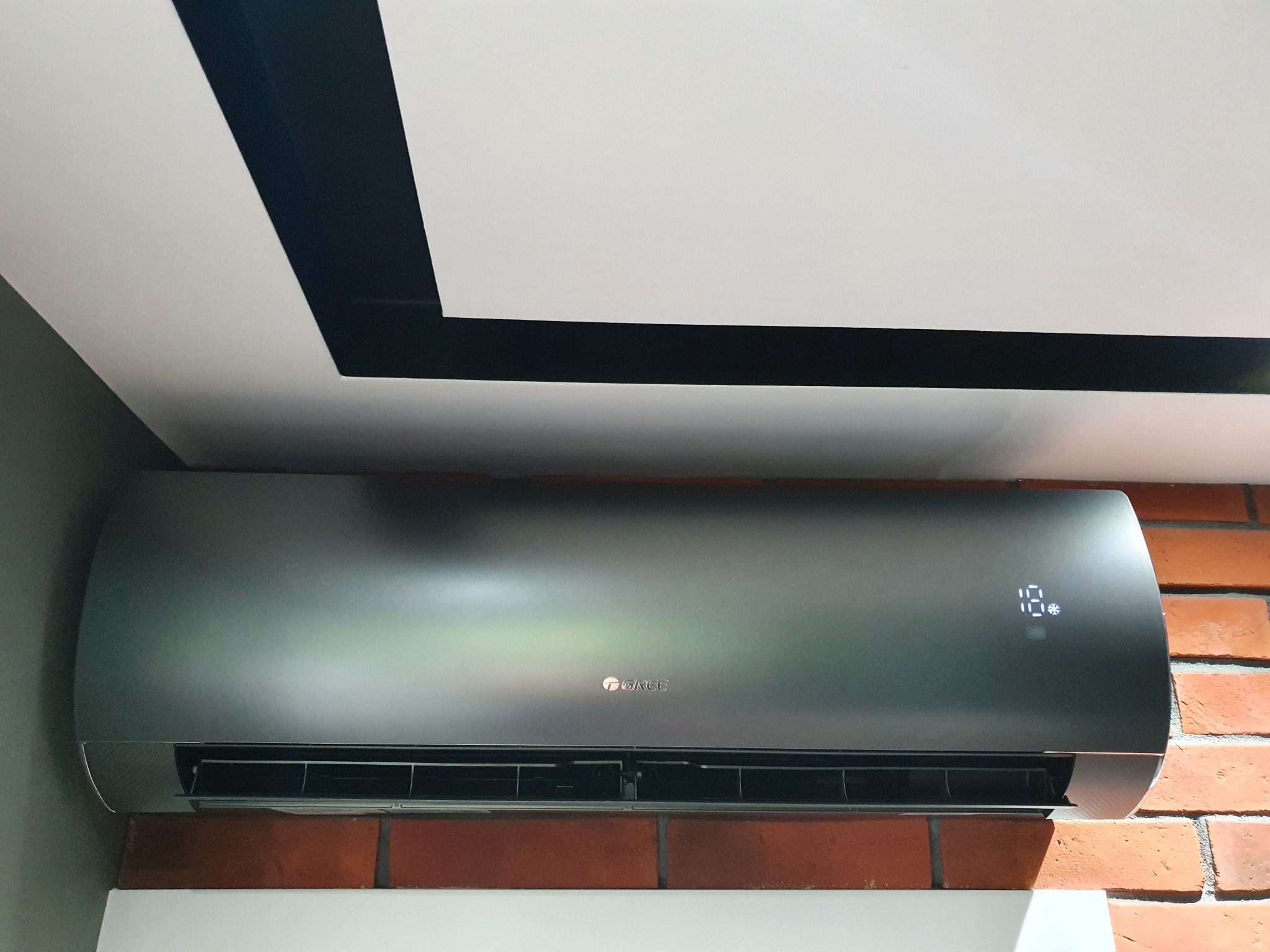 Czarny klimatyzator marki Gree zamontowany na ścianie z czerwonej cegły, widoczny wyświetlacz z temperaturą 18 stopni, biały sufit z czarnymi elementami dekoracyjnymi.