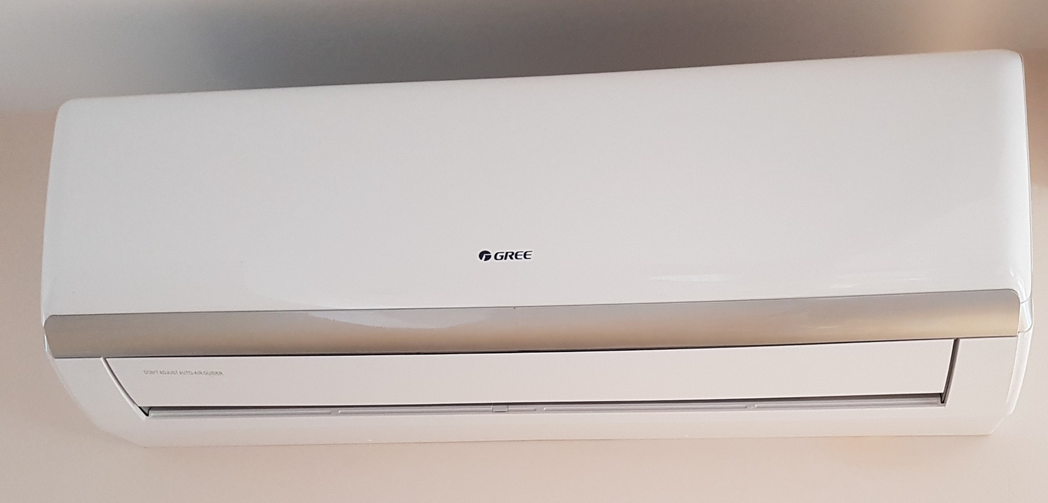 Biała jednostka wewnętrzna klimatyzatora marki Gree zamontowana na jasnej ścianie, widoczne logo producenta i informacja 'Don't adjust auto-air guider'.
