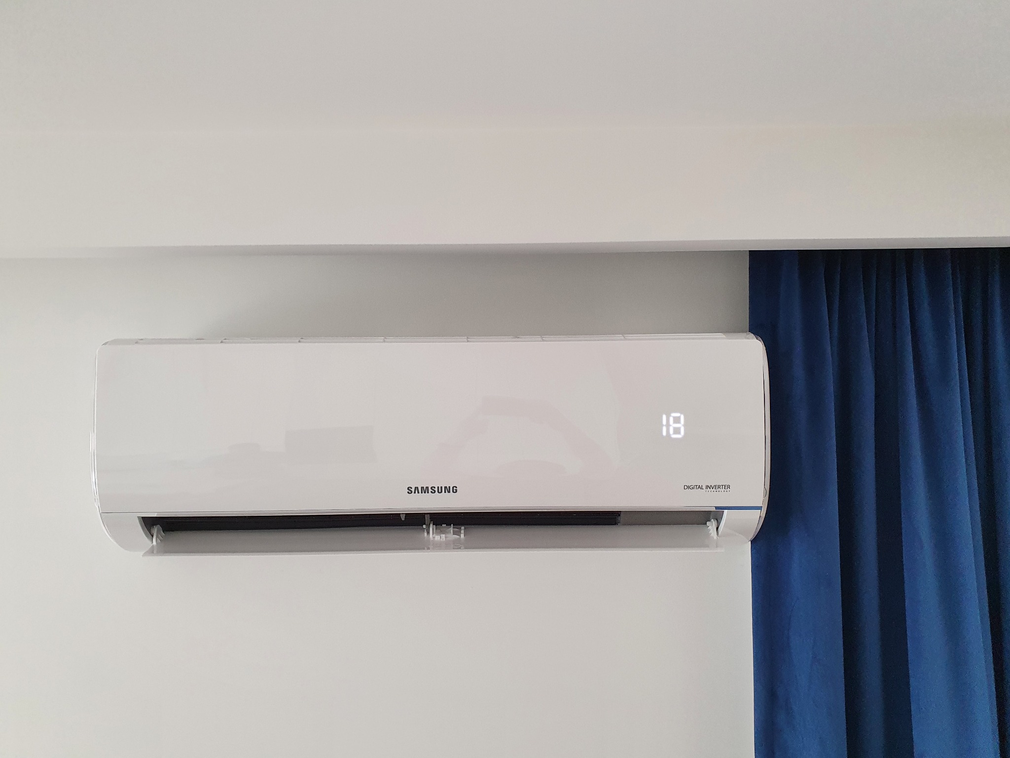 Biały klimatyzator Samsung zamontowany na ścianie, wyświetlający temperaturę 18 stopni, z fragmentem zasłony w tle.