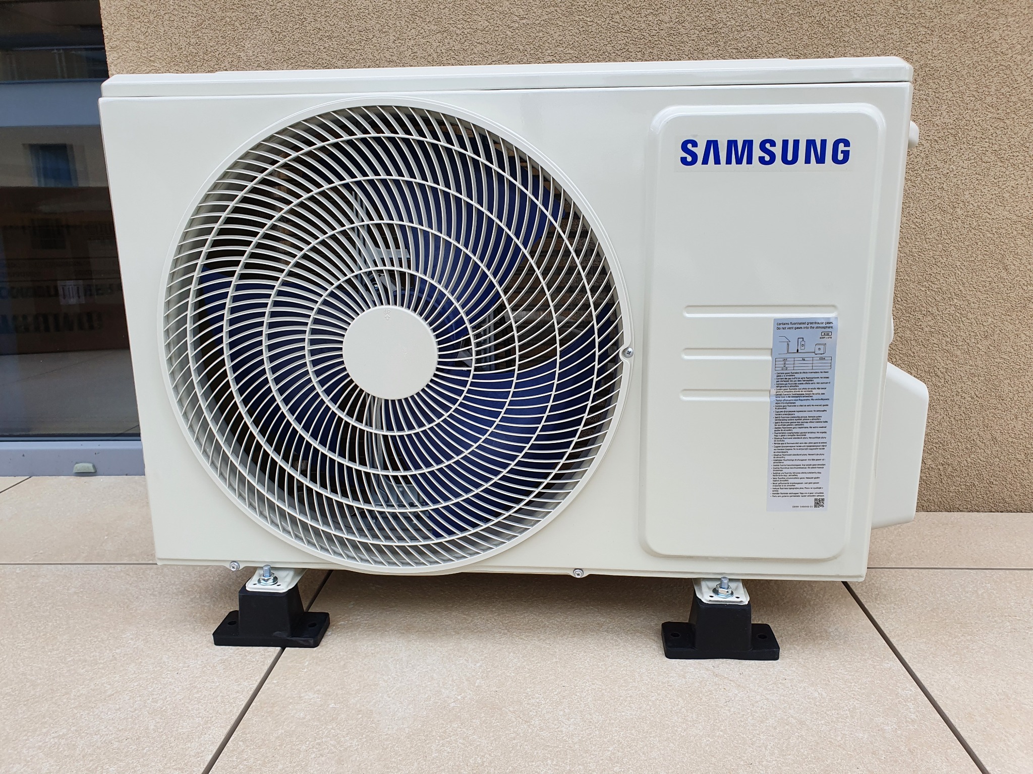Zewnętrzna jednostka klimatyzacji Samsung, zamontowana na czarnych, gumowych podstawkach na jasnobeżowej, płytkowej powierzchni, z widoczną etykietą informacyjną na obudowie.