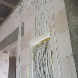 Rozprowadzenie przewodów elektrycznych w ścianie z betonu komórkowego, widoczna biała skrzynka rozdzielcza z licznymi kablami wewnątrz, kable prowadzone po ścianie mocowane uchwytami, surowy stan...