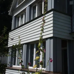 Trzykondygnacyjny dom z szarej, poziomej deski elewacyjnej, z satelitą na bocznej ścianie i malwami w pierwszym planie.