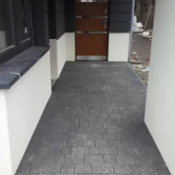 Ułożona kostka brukowa w odcieniach szarości przed wejściem do domu z brązowymi drzwiami i elementami elewacji w kolorze grafitowym.