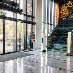 Rombud - Nowoczesne wnętrze biurowe z przeszklonymi drzwiami wejściowymi, schodami z elementami zieleni i marmurową podłogą z refleksami świetlnymi.