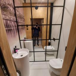 Rombud - Nowoczesna łazienka z białymi umywalkami na postumentach, toaletą podwieszaną i czarną, przeszkloną ścianką działową z widocznym odbiciem fotografa.