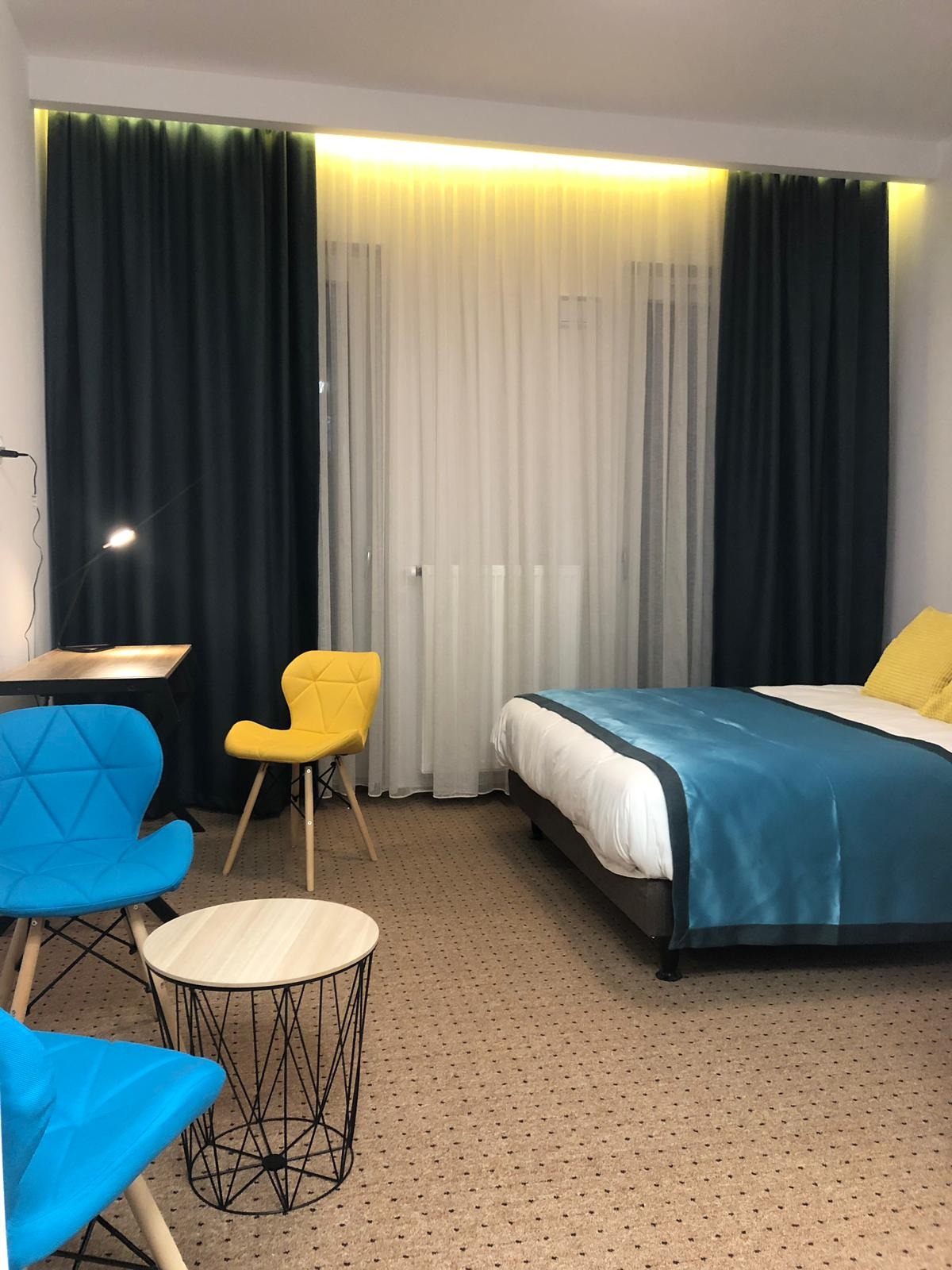 Nowoczesna sypialnia z niebieskimi i żółtymi akcentami: turkusowa narzuta na łóżku, geometryczne niebieskie krzesła, żółte krzesło przy biurku, okrągły stolik kawowy z metalową podstawą, ciemne...