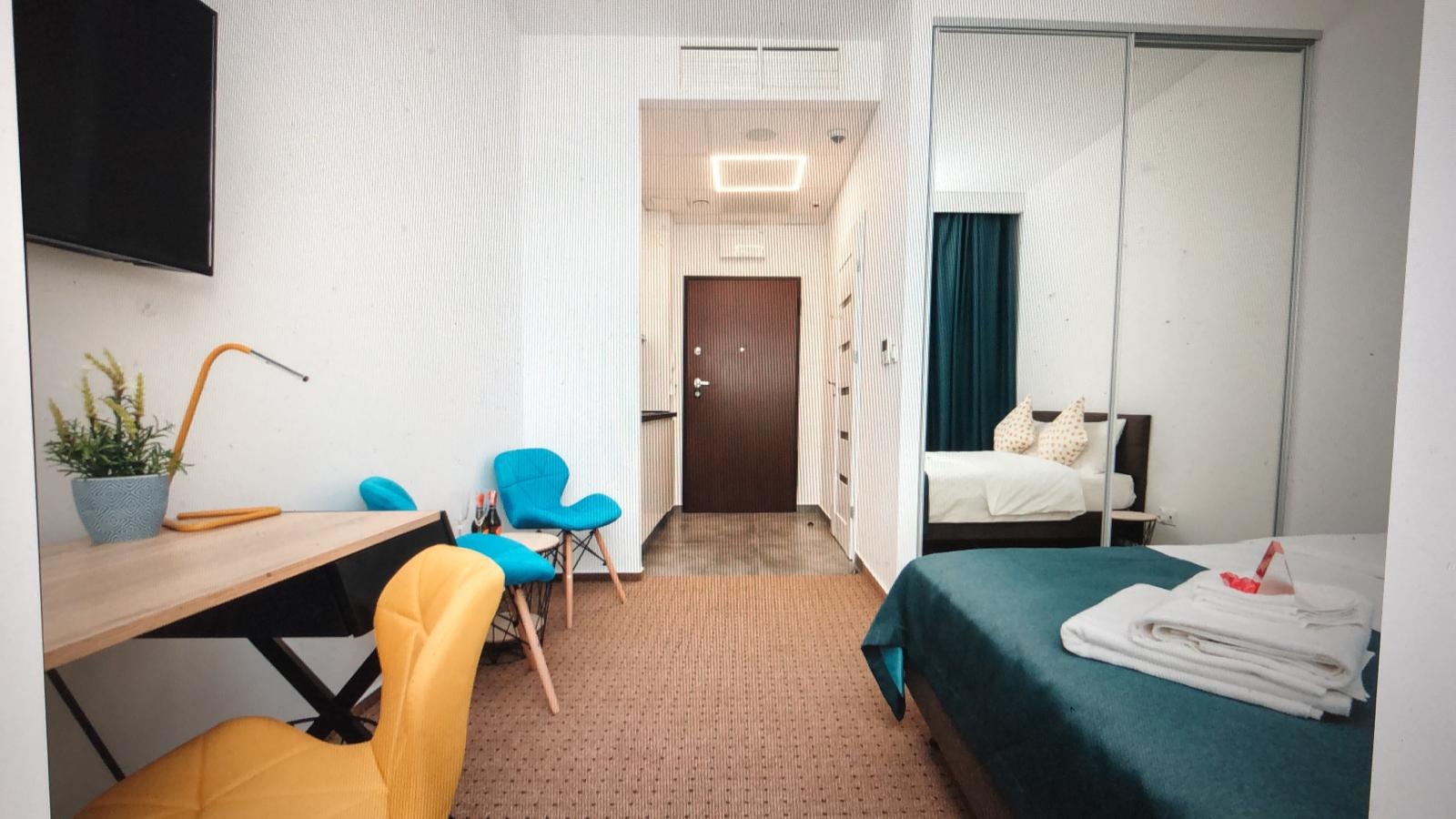 Nowoczesny pokój w Warszawie z biurkiem, lampką, turkusowymi krzesłami i łóżkiem z zieloną narzutą, widok na korytarz z drzwiami wejściowymi i szafą z lustrem.