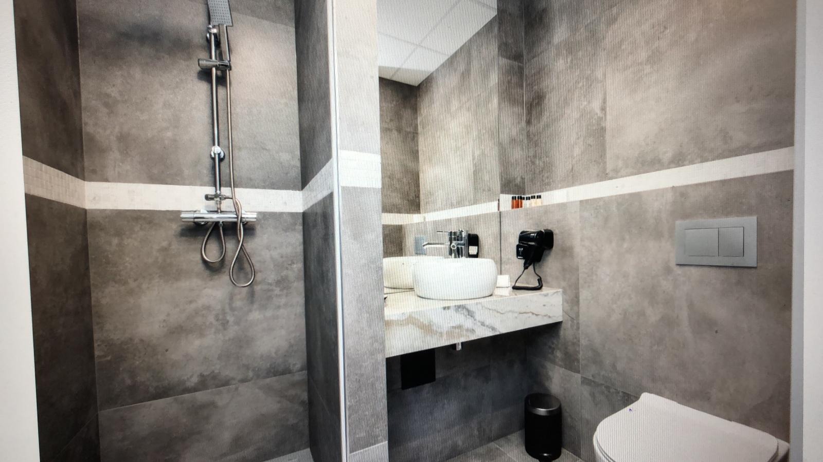 Nowoczesna łazienka z szarymi płytkami imitującymi beton, białą umywalką nablatową na marmurowym blacie, chromowaną armaturą i kabiną prysznicową typu walk-in.