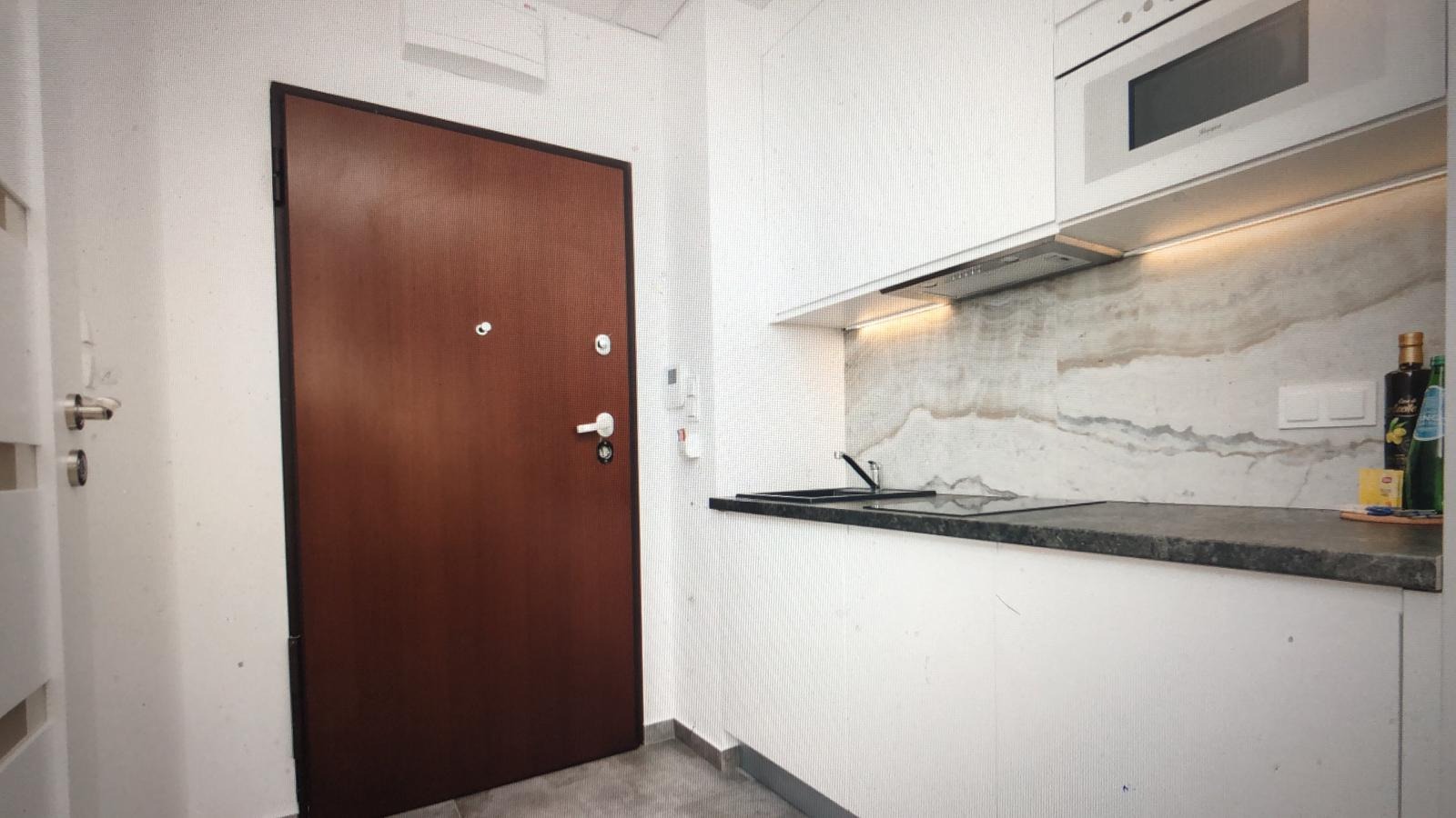 Wnętrze mieszkania z brązowymi drzwiami wejściowymi, białą zabudową kuchenną, kamiennym blatem i marmurowym panelem ściennym.