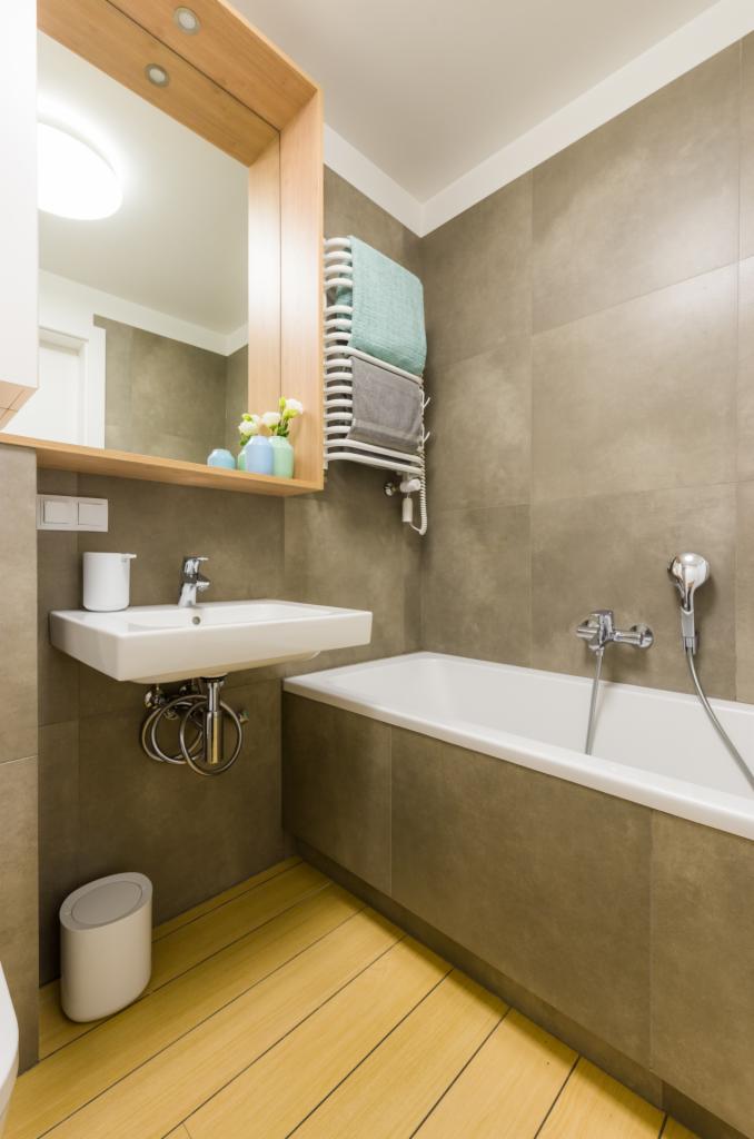 Nowoczesna łazienka z beżowymi płytkami, drewnianą podłogą, białą wanną i umywalką, oraz grzejnikiem drabinkowym z ręcznikami.