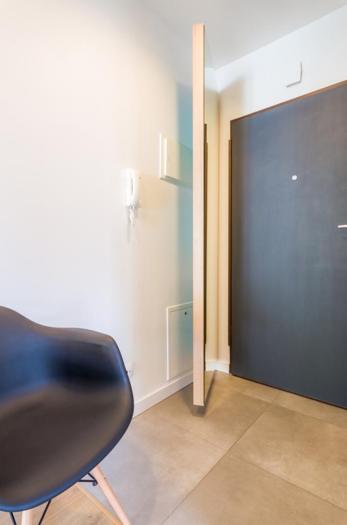 Nowoczesne lustro w drewnianej ramie, ustawione w rogu przedpokoju z szarą podłogą z płytek i czarnym krzesłem, na ścianie domofon.