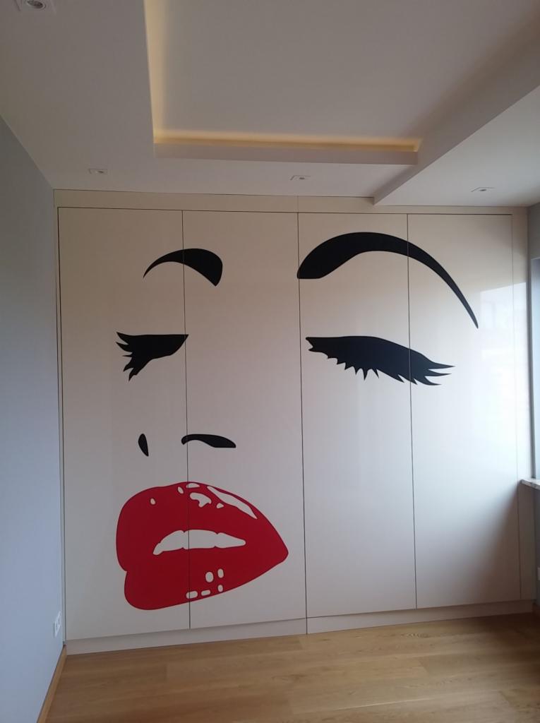 Duża grafika przedstawiająca twarz kobiety w stylu pop-art na drzwiach szafy wnękowej, z czerwonymi ustami i czarnymi brwiami i rzęsami, w jasnym wnętrzu z drewnianą podłogą.