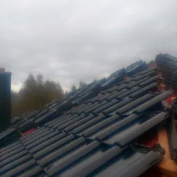 Układanie błyszczącej, czarnej dachówki ceramicznej na czerwonej membranie dachowej, widoczne drewniane łaty i stosy dachówek czekające na montaż, pochmurne niebo w tle.