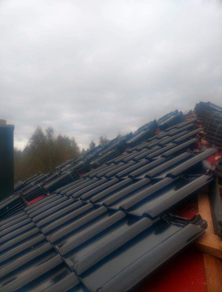 Układanie błyszczącej, czarnej dachówki ceramicznej na czerwonej membranie dachowej, widoczne drewniane łaty i stosy dachówek czekające na montaż, pochmurne niebo w tle.