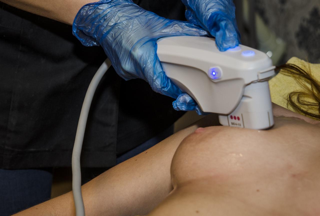 Zabieg liftingu piersi urządzeniem emitującym fale ultradźwiękowe, wykonywany przez osobę w niebieskich rękawiczkach ochronnych; widoczne włączone diody na urządzeniu.