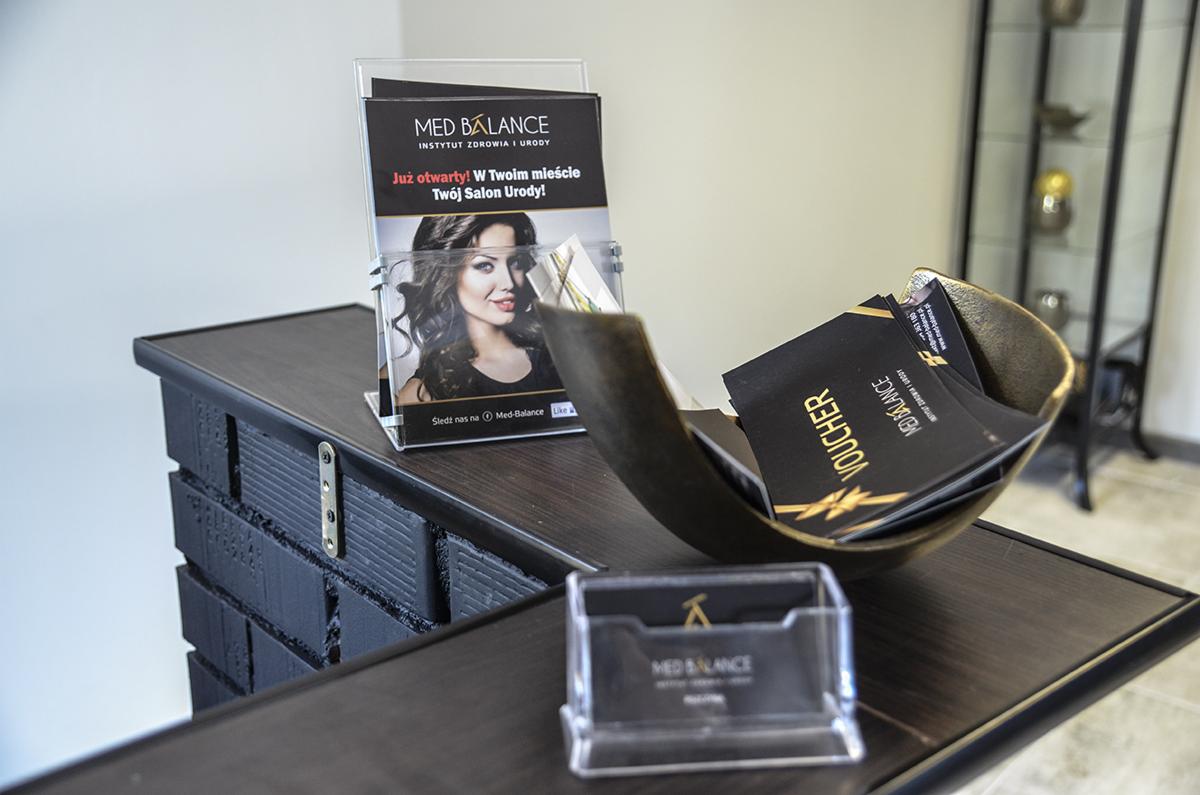 Stojak z ulotkami i wizytówkami Med Balance Instytut Zdrowia i Urody, złota miseczka z voucherami na ciemnym drewnianym blacie w salonie.