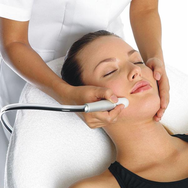 Zabieg na twarz z użyciem urządzenia emitującego fale radiowe, wykonywany przez kosmetyczkę, pacjentka z zamkniętymi oczami leży na białym ręczniku.